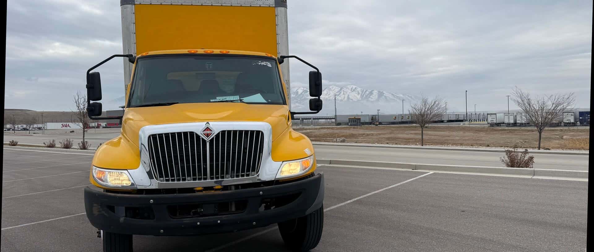 Front view of the yellow International box truck with mountains and a freight terminal behind it.