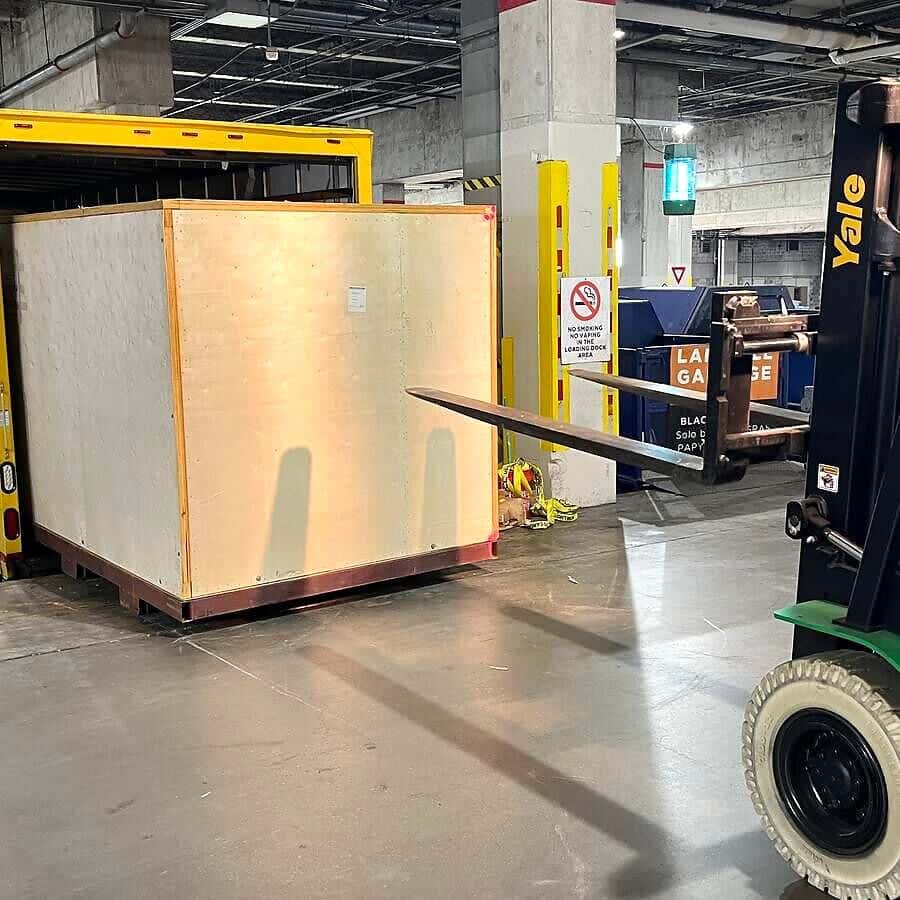 A large wooden crate is unloaded from the yellow box truck at an underground loading dock.