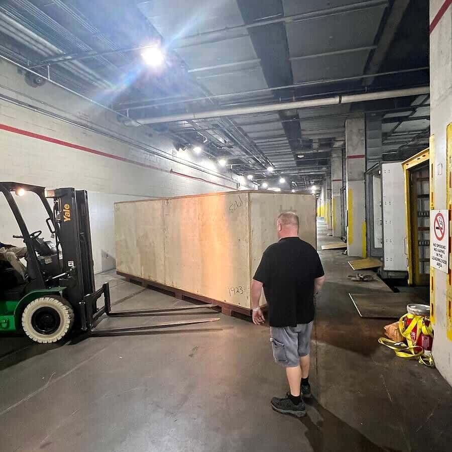 A large wooden crate on pallets in a loading dock corridor, with a forklift and a worker nearby.